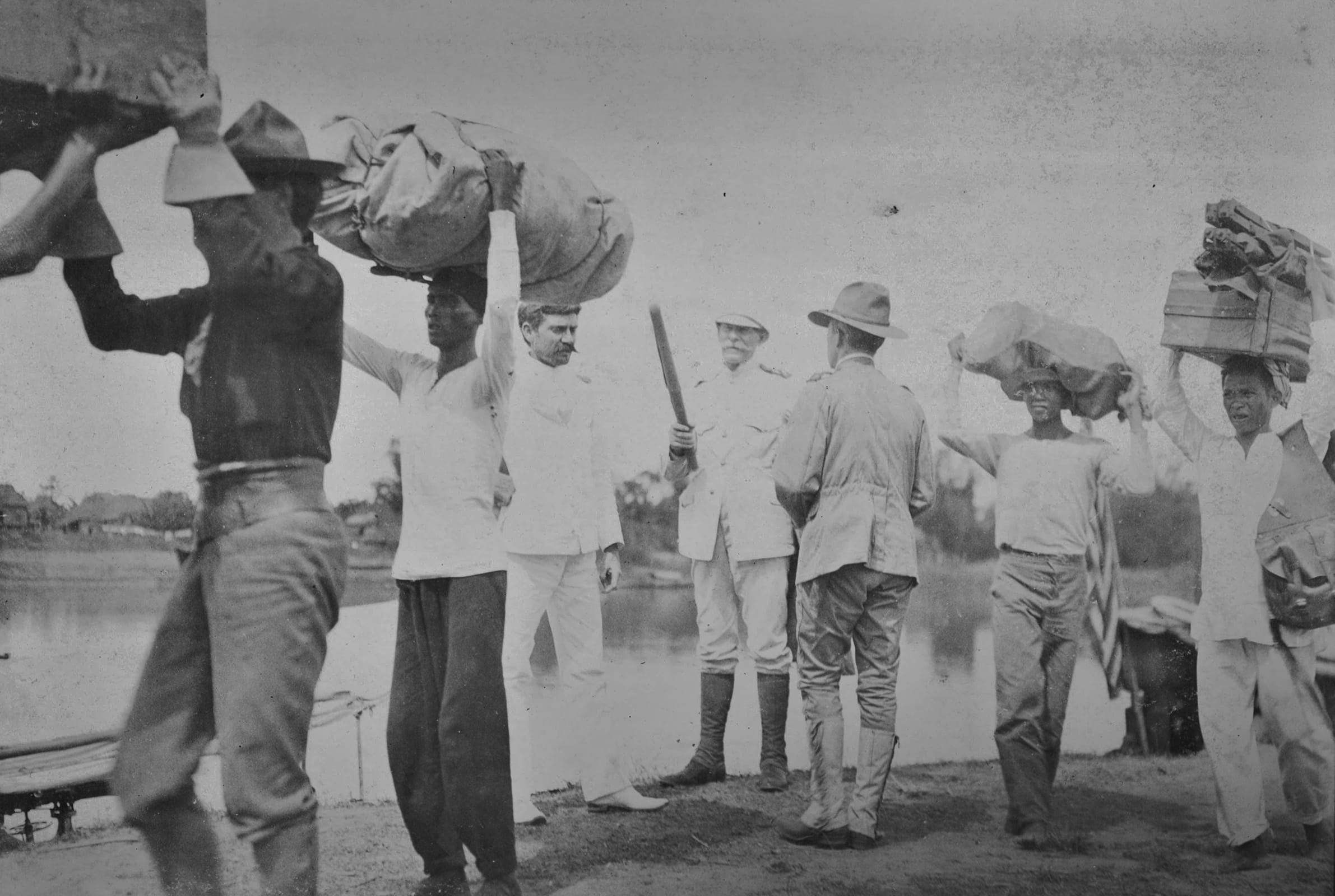 A white man in military garb stands holding a baton while several darker-skinned people walk by carrying large items.